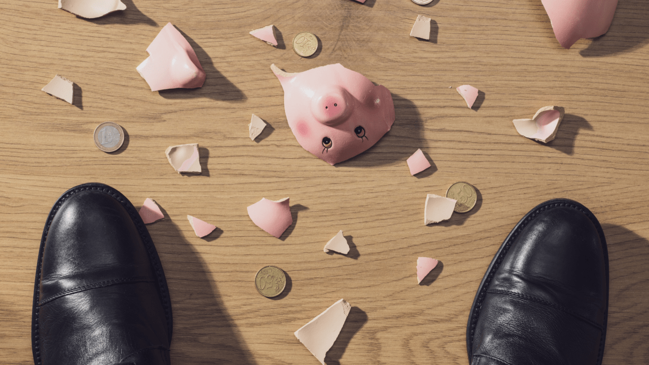 Broken Piggy bank with shards and coins all around in front of a man's formal shoes.