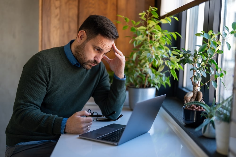 Worried man looking at laptop while holding eyeglasses, thinking about finances