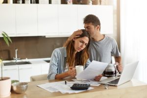 A worried couple reviewing debt documents at the kitchen table with a laptop and a calculator