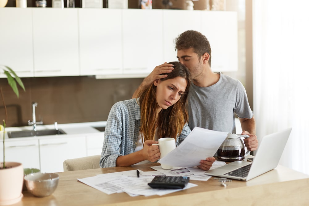 A worried couple reviewing debt documents at the kitchen table with a laptop and a calculator