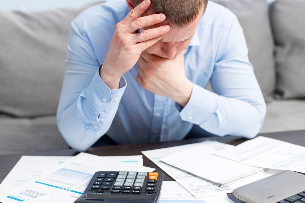 Stressed man calculating finances with bills and documents on table