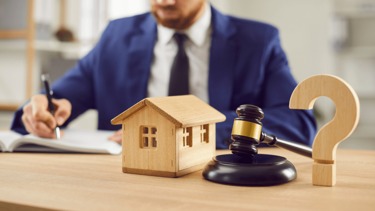 Bankruptcy attorney in blue suit working at desk with wooden house model, gavel, and question mark representing can I file for bankruptcy without my spouse legal consultation