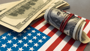 Roll of $100 bills secured with a red rubber band resting on an American flag alongside scattered currency